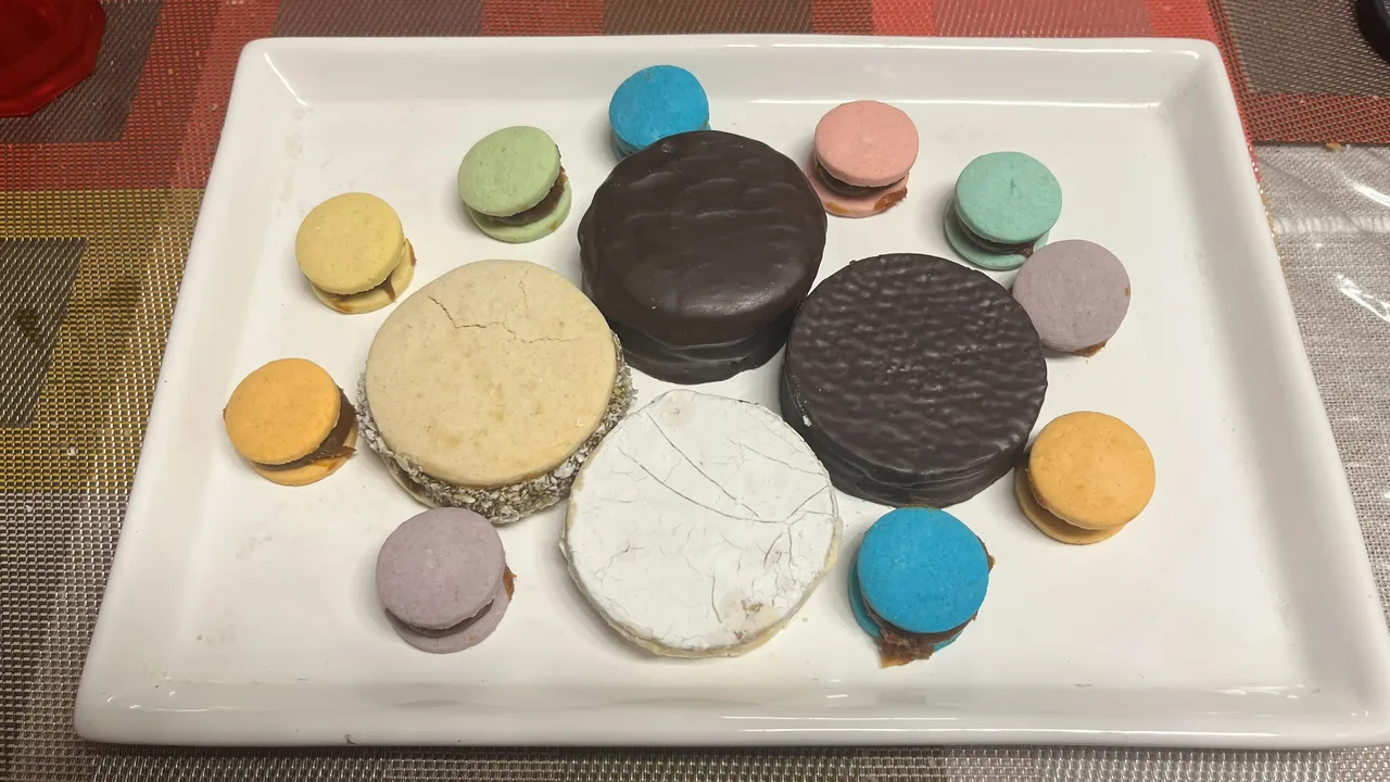 Een bord met alfajores van heel verschillende maten: één van maïzena met kokosrand, één geglazuurd, twee met chocolade omhuld en tien kleine gekleurde
