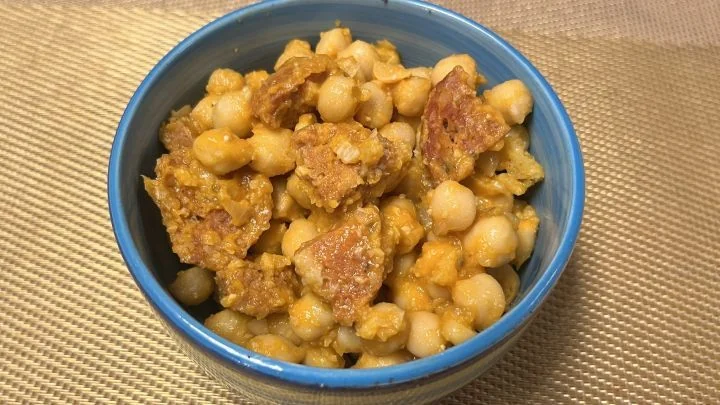 Kikhernepataa ja chorizo-makkaran paloja sinisessä kulhossa, jossa on kermainen oranssi paahdettu tomaattikastike.
