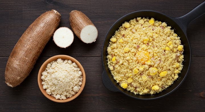 Ganze und aufgeschnittene Maniokwurzel mit weißem Fruchtfleisch, eine Holzschale mit körnigem Maniokmehl und eine gusseiserne Pfanne mit goldbrauner Farofa