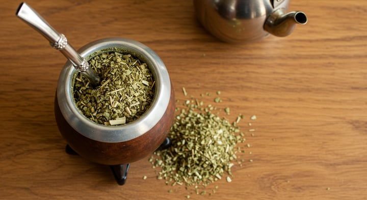 Cuia de mate com bomba cheia de erva, um montinho de erva solta ao lado mostrando folha quebrada e palito, e uma chaleira